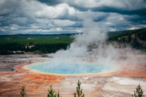 A geothermal hot spring emits steam, surrounded by colorful mineral-rich water in hues of blue, orange, and brown. In the background, a dense forest stretches under a cloudy sky, with small groups of people observing from a distance.