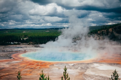 A geothermal hot spring emits steam, surrounded by colorful mineral-rich water in hues of blue, orange, and brown. In the background, a dense forest stretches under a cloudy sky, with small groups of people observing from a distance.