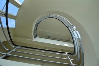 A spiral staircase viewed from below, featuring an upward curve and metal rails. The staircase appears to be indoors, with natural light coming through glass panels adjacent to the structure. The architecture is modern, with clean lines and a minimalist design.