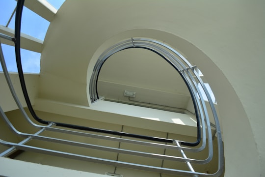 A spiral staircase viewed from below, featuring an upward curve and metal rails. The staircase appears to be indoors, with natural light coming through glass panels adjacent to the structure. The architecture is modern, with clean lines and a minimalist design.