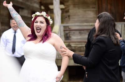 A joyful bride with vibrant pink hair and a floral crown is celebrating, with her arm raised high. She is wearing a white strapless wedding dress and is laughing exuberantly. People around her are smiling and appear to be congratulating her.