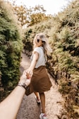 A woman with long blonde hair is walking along a path surrounded by greenery, holding hands with another person. She wears sunglasses, a white shirt, and a brown skirt, while the other person has a watch on their wrist. The scene conveys a sense of joy and adventure.