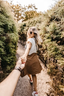 A woman with long blonde hair is walking along a path surrounded by greenery, holding hands with another person. She wears sunglasses, a white shirt, and a brown skirt, while the other person has a watch on their wrist. The scene conveys a sense of joy and adventure.
