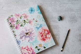 A spiral-bound notebook with a floral cover featuring large, colorful flowers rests on a light gray surface. Beside the notebook are a black pencil and a small piece of gray eraser.