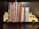 A collection of children's books neatly organized between two yellow bookends shaped like school buses, placed on a dark wooden shelf. The books have colorful spines and include popular titles, creating a bright and playful arrangement.