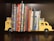 A collection of children's books neatly organized between two yellow bookends shaped like school buses, placed on a dark wooden shelf. The books have colorful spines and include popular titles, creating a bright and playful arrangement.