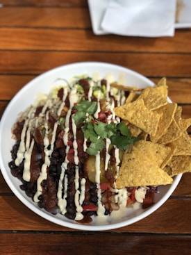 A white bowl filled with a colorful assortment of food sits on a wooden table. The dish features black beans, diced tomatoes, pickled jalapeños, and is drizzled with a creamy white sauce. Fresh cilantro is garnished on top, and there are tortilla chips on one side of the bowl.