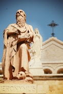 A large stone statue of a robed, bearded man holding a quill and a book. At the base of the statue, there's a skull. In the background, there is another statue of a robed figure and a cross atop a building with a light beige facade and arches.