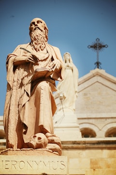A large stone statue of a robed, bearded man holding a quill and a book. At the base of the statue, there's a skull. In the background, there is another statue of a robed figure and a cross atop a building with a light beige facade and arches.