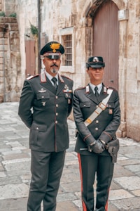 Two uniformed individuals stand side by side on a cobblestone street lined with historic stone buildings. They wear dark military-style uniforms adorned with medals and insignia. One person has a mustache and wears a peaked cap with an emblem, and the other wears a similar hat and gloves.