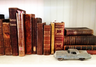 Several vintage books with worn, ornate covers are lined up on a shelf. In front of the books, a small, blue toy car is placed, adding a whimsical contrast to the historical feel of the books. The setting suggests a cozy, nostalgic atmosphere.