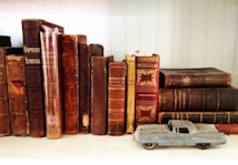 Several vintage books with worn, ornate covers are lined up on a shelf. In front of the books, a small, blue toy car is placed, adding a whimsical contrast to the historical feel of the books. The setting suggests a cozy, nostalgic atmosphere.