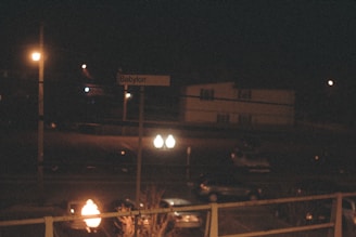 A nighttime scene featuring a dark street illuminated by a few streetlights. A sign reading 'Babylon' is visible, with a residential building and parked cars in the background. The scene is dimly lit, creating an atmospheric mood.