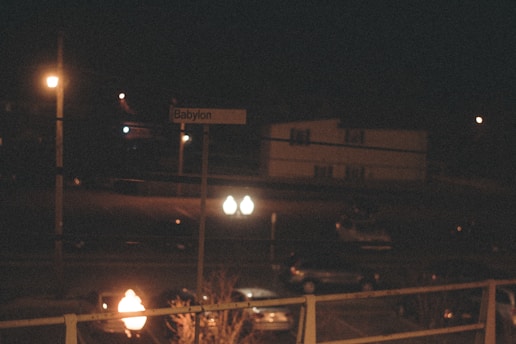 A nighttime scene featuring a dark street illuminated by a few streetlights. A sign reading 'Babylon' is visible, with a residential building and parked cars in the background. The scene is dimly lit, creating an atmospheric mood.