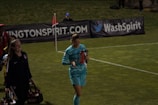 Two individuals walk along a grassy field, one wearing a teal sports outfit holding a red water bottle. A third person in the background stands near advertising banners. The scene appears to be at a sports event, likely a soccer match judging by the field lines and corner flag.