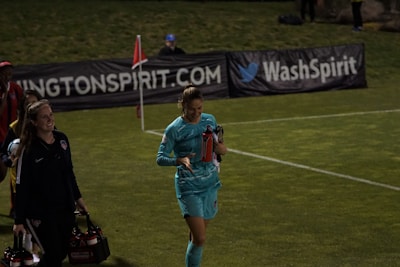 Two individuals walk along a grassy field, one wearing a teal sports outfit holding a red water bottle. A third person in the background stands near advertising banners. The scene appears to be at a sports event, likely a soccer match judging by the field lines and corner flag.