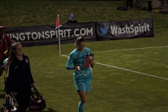Two individuals walk along a grassy field, one wearing a teal sports outfit holding a red water bottle. A third person in the background stands near advertising banners. The scene appears to be at a sports event, likely a soccer match judging by the field lines and corner flag.