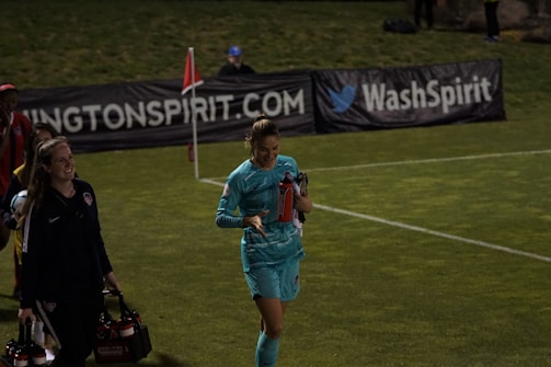 Two individuals walk along a grassy field, one wearing a teal sports outfit holding a red water bottle. A third person in the background stands near advertising banners. The scene appears to be at a sports event, likely a soccer match judging by the field lines and corner flag.