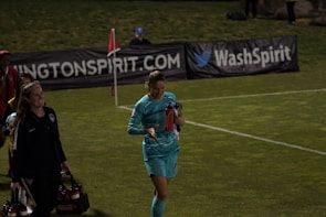 Two individuals walk along a grassy field, one wearing a teal sports outfit holding a red water bottle. A third person in the background stands near advertising banners. The scene appears to be at a sports event, likely a soccer match judging by the field lines and corner flag.