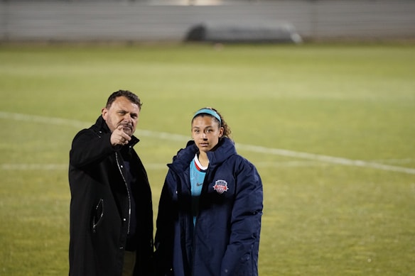 A man and a woman are standing on a sports field. The man, dressed in a dark coat, is gesturing forward with his hand while the woman, wearing a blue sports jacket with a logo, looks on. The background features a grassy field with faint lines visible on the ground.