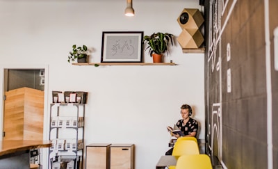 A cozy interior with a modern aesthetic, featuring a person sitting and reading a book beside a chalkboard wall with drawings. There are yellow chairs, a shelf with plants and framed art, and wooden storage boxes.