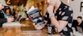 A person in a floral shirt is sitting at a wooden table reading a book titled 'Expert Secrets' by Russell Brunson. Next to them is a laptop and a black mug with '80/20' printed on it. The background features a blurred cafe environment with other people, creating a relaxed and cozy atmosphere.