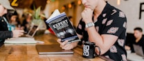 A person in a floral shirt is sitting at a wooden table reading a book titled 'Expert Secrets' by Russell Brunson. Next to them is a laptop and a black mug with '80/20' printed on it. The background features a blurred cafe environment with other people, creating a relaxed and cozy atmosphere.