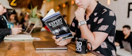 A person in a floral shirt is sitting at a wooden table reading a book titled 'Expert Secrets' by Russell Brunson. Next to them is a laptop and a black mug with '80/20' printed on it. The background features a blurred cafe environment with other people, creating a relaxed and cozy atmosphere.