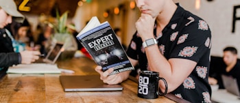 A person in a floral shirt is sitting at a wooden table reading a book titled 'Expert Secrets' by Russell Brunson. Next to them is a laptop and a black mug with '80/20' printed on it. The background features a blurred cafe environment with other people, creating a relaxed and cozy atmosphere.