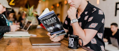 A person in a floral shirt is sitting at a wooden table reading a book titled 'Expert Secrets' by Russell Brunson. Next to them is a laptop and a black mug with '80/20' printed on it. The background features a blurred cafe environment with other people, creating a relaxed and cozy atmosphere.