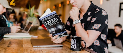 A person in a floral shirt is sitting at a wooden table reading a book titled 'Expert Secrets' by Russell Brunson. Next to them is a laptop and a black mug with '80/20' printed on it. The background features a blurred cafe environment with other people, creating a relaxed and cozy atmosphere.