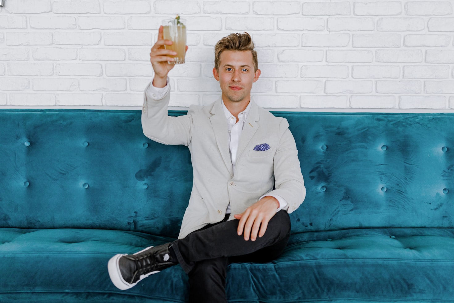 Blonde man sitting on blue couch wearing grey blazer holds up his cocktail while looking at the camera.