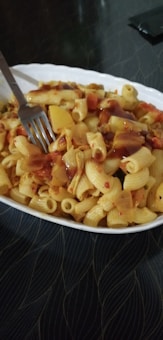 A bowl filled with pasta, mixed with a variety of vegetables, is topped with a brown tomato-based sauce. A fork is placed in the bowl, indicating it is ready to be eaten. The surface underneath the bowl has a subtle wavy pattern.