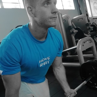 A man is working out in a gym, wearing a bright blue t-shirt with the words 'MOVE SMRT' on it. He is using a barbell for his exercise and appears to be focused and determined. There is gym equipment visible in the background.