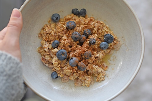 A hand holds a bowl containing oatmeal topped with fresh blueberries and a sprinkle of cinnamon. The oatmeal appears creamy, suggesting it might be mixed with milk or water.