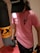 A person wearing a pink T-shirt and a black cap is holding a dumbbell in one hand. They are standing next to a punching bag, suggesting a fitness or gym setting.