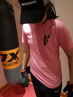 A person wearing a pink T-shirt and a black cap is holding a dumbbell in one hand. They are standing next to a punching bag, suggesting a fitness or gym setting.