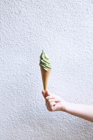 A hand holds a cone of soft serve ice cream with a pale green color against a textured white wall background.