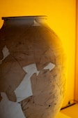 A large, pottery vase is visible, showcasing a patchwork of repaired sections. The surface displays several cracks and uneven textures, highlighting its aged and restored condition. The background is warmly lit, casting an amber glow.