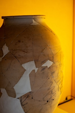 A large, pottery vase is visible, showcasing a patchwork of repaired sections. The surface displays several cracks and uneven textures, highlighting its aged and restored condition. The background is warmly lit, casting an amber glow.