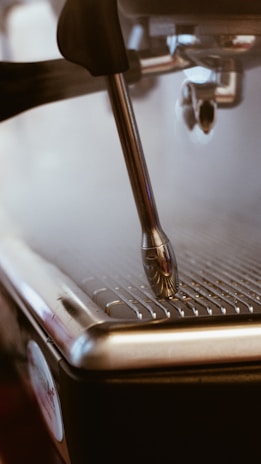 Close-up view of a coffee machine's steam wand, with steam rising as the metal nozzle hovers over a metal grate. The focus is on the metallic components, with soft lighting creating a warm and inviting atmosphere.