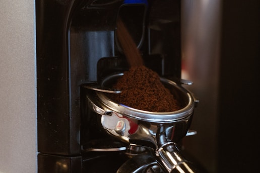 Freshly ground coffee is being dispensed into a portafilter from a coffee grinder. The machine is metallic and has a sleek, modern design.