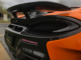 A close-up view of the rear section of an orange sports car, featuring a prominent black carbon fiber rear wing and sleek taillights. The car's aerodynamic design is evident, with smooth curves and a glossy finish.