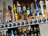 A row of various beer taps is mounted above a selection of bottled beers. The taps feature diverse and colorful designs, including a pirate figure, a gnome, and different logos. The background consists of a blurred brick wall and signage.