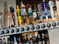 A row of various beer taps is mounted above a selection of bottled beers. The taps feature diverse and colorful designs, including a pirate figure, a gnome, and different logos. The background consists of a blurred brick wall and signage.