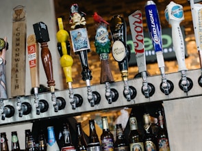 A row of various beer taps is mounted above a selection of bottled beers. The taps feature diverse and colorful designs, including a pirate figure, a gnome, and different logos. The background consists of a blurred brick wall and signage.
