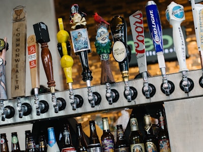 A row of various beer taps is mounted above a selection of bottled beers. The taps feature diverse and colorful designs, including a pirate figure, a gnome, and different logos. The background consists of a blurred brick wall and signage.