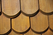 Close-up of a wooden shingle roof with overlapping, semi-circular tiles displaying a natural wood grain pattern. The shingles are arranged in a uniform pattern, indicating craftsmanship and durability.