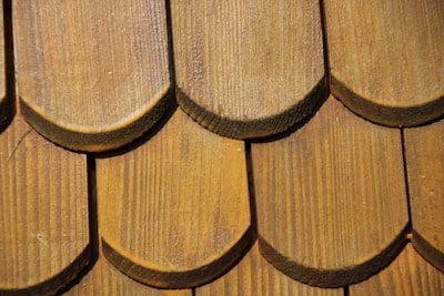 Close-up of a wooden shingle roof with overlapping, semi-circular tiles displaying a natural wood grain pattern. The shingles are arranged in a uniform pattern, indicating craftsmanship and durability.