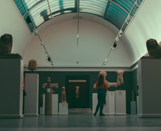 A person stands in an art gallery or museum hall observing sculptures that are displayed on pedestals. The sculptures appear to be ancient torso statues and heads made from stone. The hall has a high arched ceiling with skylights, and the lighting is subdued.